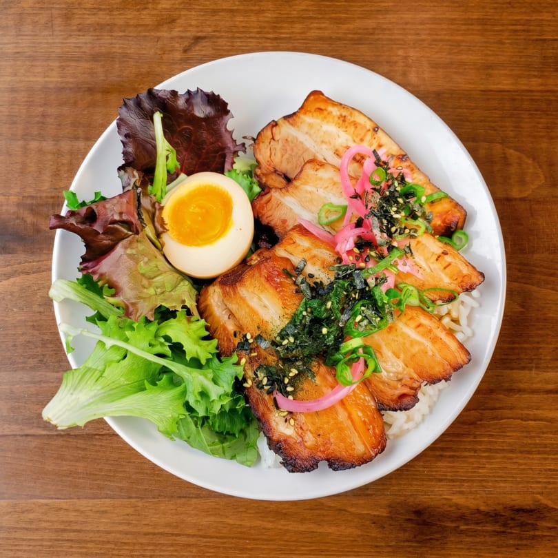 A white bowl of chashu pork, rice, soft-boiled egg, mixed greens, and pickled onions.