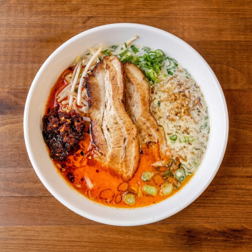 Overhead view of a ramen bowl with pork belly, bean sprouts, chili oil, and green onions.
